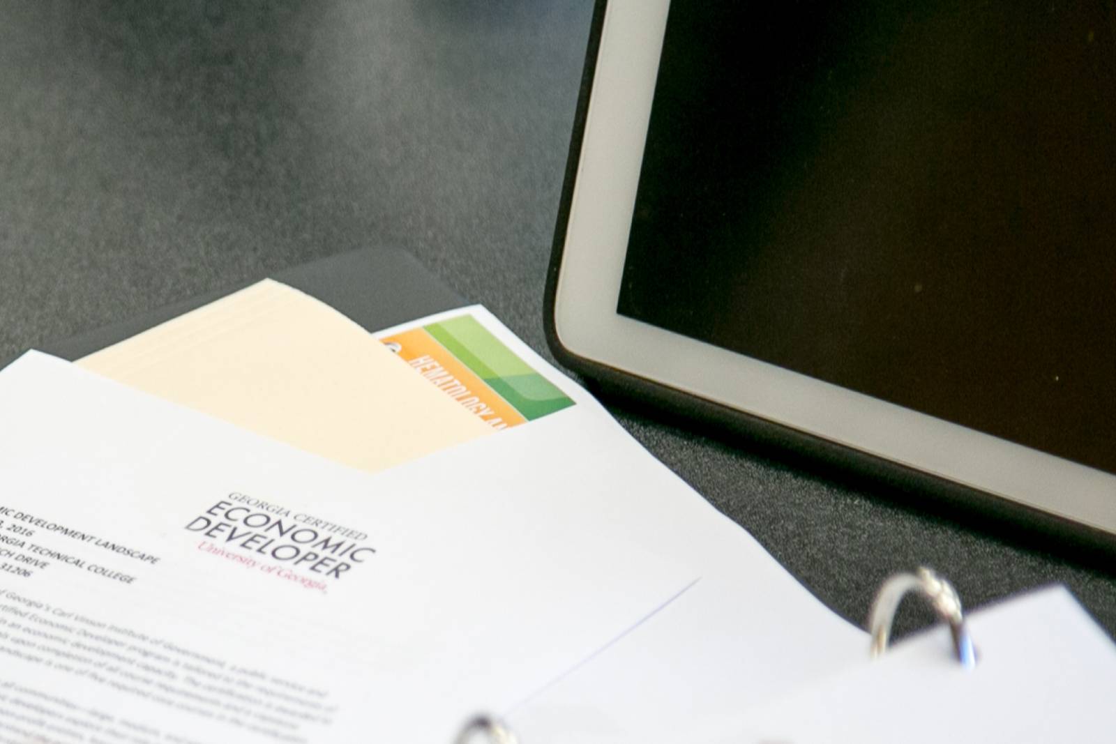 Close-up of a binder with a document labeled “Georgia Certified Economic Developer” beside a folder and a tablet on a dark tabletop.
